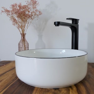 Könnte beinhalten: Ein weißes Keramik-Waschbecken mit schwarzem Rand und einem schwarzen Wasserhahn. Eine rosa Vase mit getrockneten Blumen befindet sich im Hintergrund. Das Waschbecken steht auf einer Holzoberfläche.