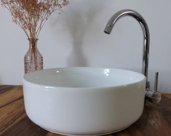 Op de afbeelding: Een witte, ronde keramische wastafel met een chromen kraan. De wastafel staat op een houten oppervlak. Een roze glazen vaas met gedroogde bloemen staat op de achtergrond.