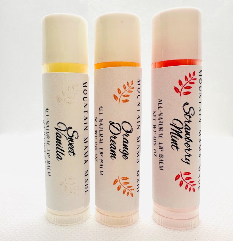 Op de afbeelding: Drie lippenbalsem tubes met witte etiketten en cr&egrave;mekleurige doppen. De etiketten lezen "Sweet Vanilla", "Orange Dream" en "Strawberry Mint". Elke tube heeft de tekst "Mountain Mama Made" en "All Natural Lip Balm".