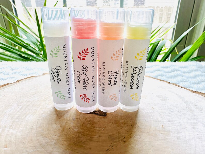 Op de afbeelding: Vier lippenbalsems met doorzichtige tubes en witte etiketten. Smaakvarianten zijn Vanille Munt, Red Velvet Cake, Fruity Cereal en Pineapple Paradise. De etiketten tonen de tekst "Mountain Mama Made" en "All Natural Lip Balm".