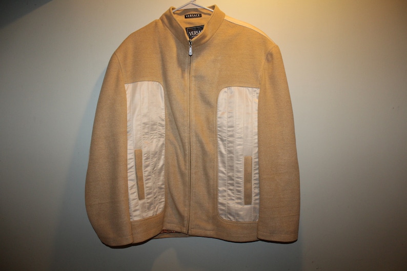 May include: A tan zip-up jacket with white panels on the sides. The jacket has a stand-up collar and the label "VERSACE" is visible on the collar.