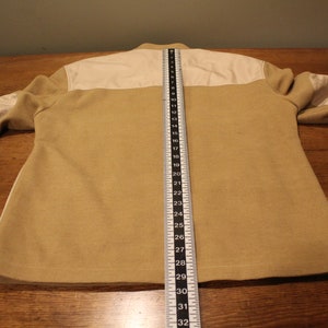May include: A beige jacket with white panels on the shoulders and elbows. The jacket is laid flat on a wooden surface with a measuring tape beside it. The tape measures 32 inches.