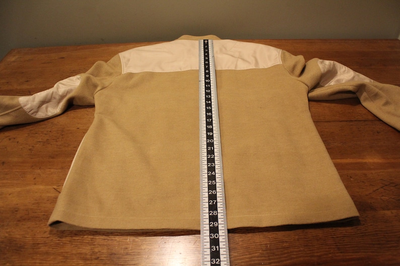 May include: A beige jacket with white panels on the shoulders and elbows. The jacket is laid flat on a wooden surface with a measuring tape beside it. The tape measures 32 inches.