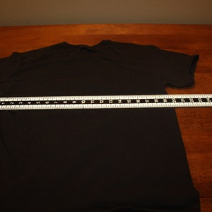 May include: A black t-shirt laid flat on a wooden surface with a measuring tape across the middle. The tape measures 50.8 cm.