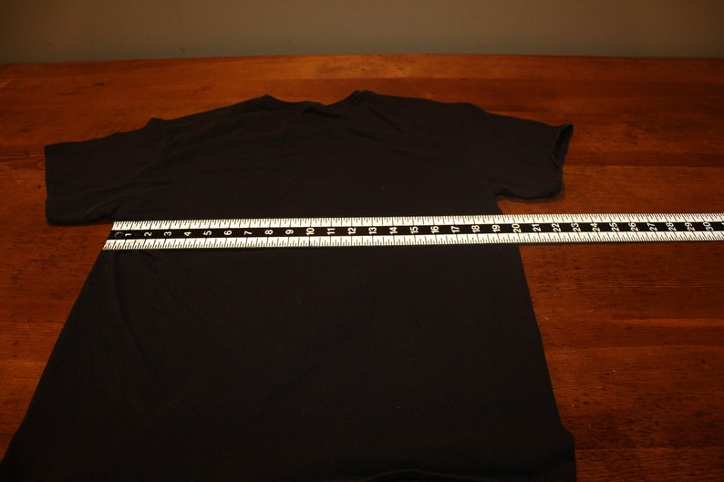 May include: A black t-shirt laid flat on a wooden surface with a measuring tape across the middle. The tape measures 50.8 cm.