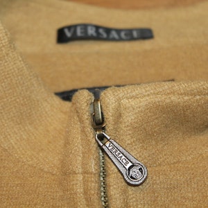 May include: A close-up of a brown zipper with a silver zipper pull. The zipper pull has the word "VERSACE" engraved on it.