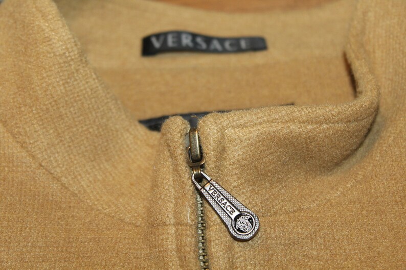 May include: A close-up of a brown zipper with a silver zipper pull. The zipper pull has the word "VERSACE" engraved on it.