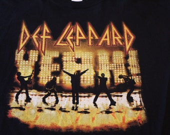 Puede incluir: Camiseta negra con un gráfico de la banda Def Leppard frente a un escenario con luces. El nombre de la banda está en letras grandes y en negrita sobre los miembros de la banda. El texto "Yeah" está en letras más pequeñas debajo del nombre de la banda.
