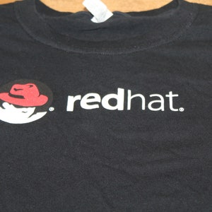 May include: Black t-shirt featuring the "red hat" logo. The logo includes a silhouette of a person wearing a red hat, with the text "red hat" in white. The shirt is made of a soft material.