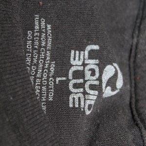May include: A black t-shirt tag with white text that reads "Liquid Blue" and "L" for size. The tag also includes care instructions: "Machine wash cold with like colours", "Only non-chlorine bleach", "Tumble dry low", "Do not dry clean", and "100% Cotton".