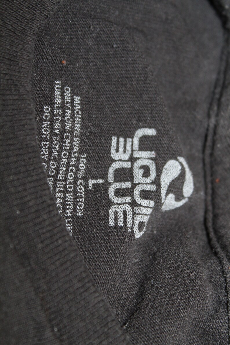 May include: A black t-shirt tag with white text that reads "Liquid Blue" and "L" for size. The tag also includes care instructions: "Machine wash cold with like colours", "Only non-chlorine bleach", "Tumble dry low", "Do not dry clean", and "100% Cotton".