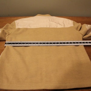 May include: A beige and white long-sleeved jacket with a white collar and a zipper closure. The jacket is laid flat on a wooden surface with a ruler measuring 25 inches across the back.