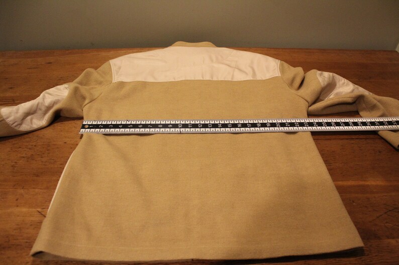 May include: A beige and white long-sleeved jacket with a white collar and a zipper closure. The jacket is laid flat on a wooden surface with a ruler measuring 25 inches across the back.