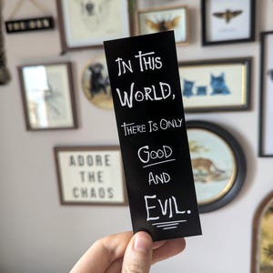 May include: A black bookmark with white text that reads "In this world, there is only good and evil." The bookmark is held in front of a wall decorated with framed art and decorative items.