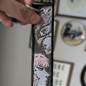 Peut inclure: Porte-clés brodé avec une illustration détaillée d'un personnage aux cheveux roses et une vue de profil. Le motif est sur un fond gris avec une bordure noire, attaché à un porte-clés argenté. Le porte-clés est rectangulaire et semble être en tissu.