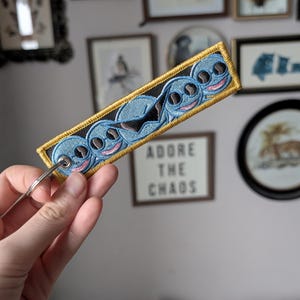 May include: Embroidered keychain with a blue alien design, pink accents, and a gold border. It has a silver ring. The text "ADORE THE CHAOS" is visible in the background. The background includes framed artwork.