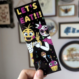 May include: A black bookmark with a colorful design featuring two cartoon characters, a yellow chicken and a pink and white chicken, with the text "Let's Eat!!!".