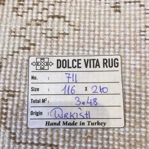 May include: A white rug with a brown and pink pattern. The rug has a label that says "DOLCE VITA RUG" and includes the following information: No. 711, Size 116 x 210, Total M&sup2; 3.48, Origin URKISH, Hand Made in Turkey.