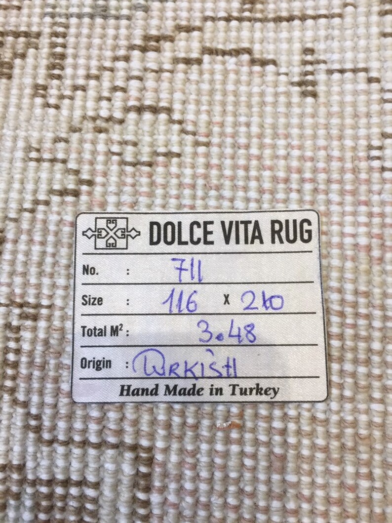 May include: A white rug with a brown and pink pattern. The rug has a label that says "DOLCE VITA RUG" and includes the following information: No. 711, Size 116 x 210, Total M&sup2; 3.48, Origin URKISH, Hand Made in Turkey.