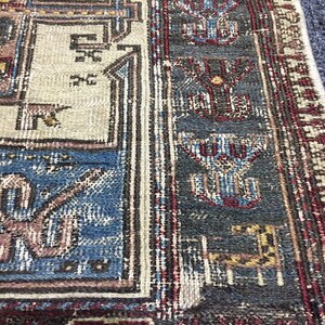 May include: Close-up of a rug with a detailed geometric pattern in shades of blue, red, brown, and white. The rug has a fringed border.