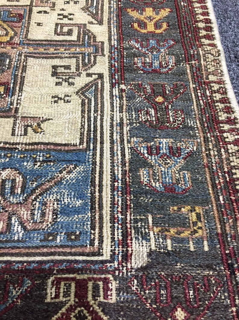May include: Close-up of a rug with a detailed geometric pattern in shades of blue, red, brown, and white. The rug has a fringed border.