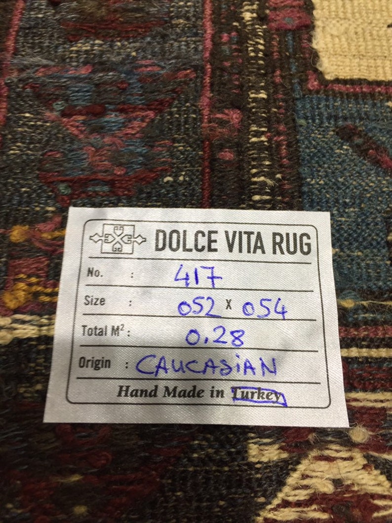 May include: A white tag with black text and blue handwritten text. The tag reads "DOLCE VITA RUG" and includes information about the rug, such as the number, size, total square metres, origin, and that it is hand-made in Turkey. The tag is on a dark brown and blue patterned rug.