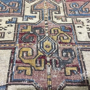 May include: Close-up of a vintage rug with a detailed geometric pattern in shades of red, blue, yellow, and beige. The rug has a worn and faded look, suggesting it is antique.
