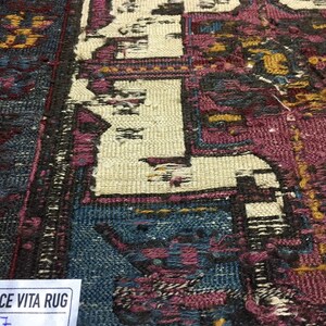 May include: Close-up of a vintage rug with a faded, intricate design in shades of blue, pink, and white. The rug has a worn, distressed look, with some areas showing signs of wear and tear. A white tag with the text "CE VITA RUG" is visible in the bottom left corner.