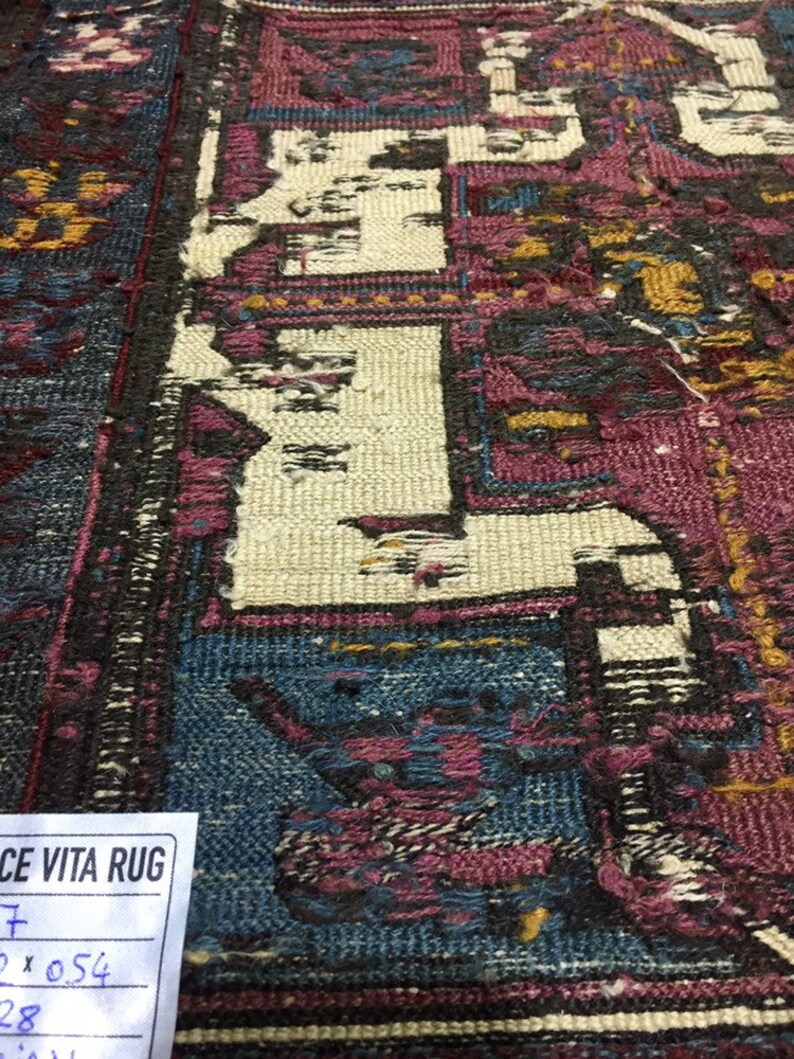 May include: Close-up of a vintage rug with a faded, intricate design in shades of blue, pink, and white. The rug has a worn, distressed look, with some areas showing signs of wear and tear. A white tag with the text "CE VITA RUG" is visible in the bottom left corner.