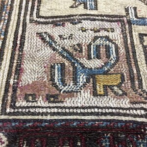 May include: Close-up of a woven rug with intricate patterns in shades of blue, brown, and white. The rug features a geometric design with a central motif of a stylised animal or bird.
