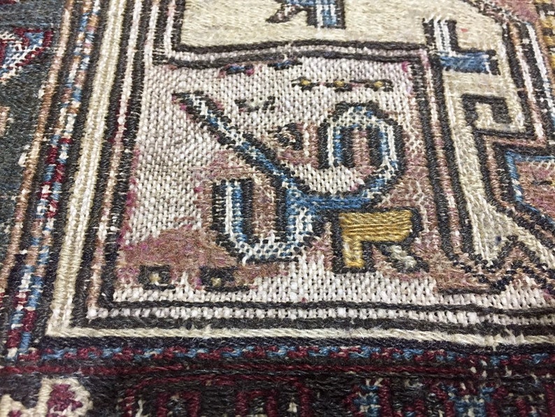 May include: Close-up of a woven rug with intricate patterns in shades of blue, brown, and white. The rug features a geometric design with a central motif of a stylised animal or bird.