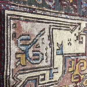 May include: Close-up of a vintage rug with intricate patterns in shades of blue, red, brown, and gold. The rug has a worn and faded appearance, suggesting it is an antique.