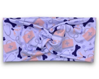 May include: A purple and pink patterned headband with a design of dog bones, first aid kits, and syringes. The headband is made of a soft, stretchy fabric.