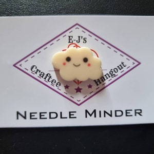 May include: A cream cloud-shaped needle minder with a smiling face. The needle minder is on a white card with the text "E-J's Craftee Hangout" inside a purple diamond and "NEEDLE MINDER" below.
