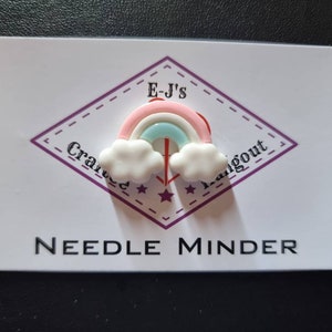 May include: A needle minder featuring a rainbow design. The rainbow is pink, blue, and red, with white clouds. The needle minder is on a white card with the text "E-J's Crafted Hangout" and "NEEDLE MINDER".