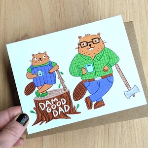 May include: A card with a cartoon illustration of two beavers. The larger beaver is wearing glasses and a green and blue plaid shirt. The smaller beaver is wearing a blue overall. The beavers are standing on a tree stump with the text "Dam Good Dad" written on it.