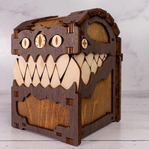 May include: A wooden box with a monster-like design. The box is made of dark brown wood with light brown teeth and eye details. The box has a hinged lid and is designed to look like a monster's face.