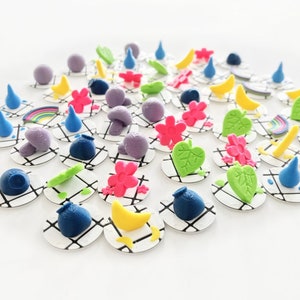 May include: A collection of colorful, miniature, 3D printed charms on white circular bases. The charms include mushrooms, rainbows, leaves, flowers, moons, and teardrops.