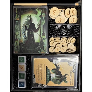 Puede incluir: Un organizador de juegos de mesa con varios componentes. Incluye cartas con ilustraciones y texto, fichas de madera con números, un dado negro y piezas de juego de colores. La caja del juego presenta una temática de fantasía.