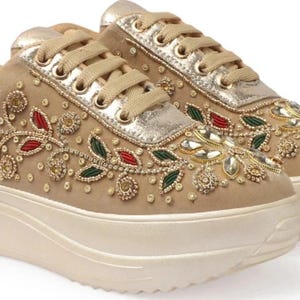 Può includere: Sneakers beige con plateau e ricami floreali intricati. Le sneakers presentano dettagli oro, rossi e verdi, con una linguetta dorata metallica e lacci abbinati. La suola platform è color beige crema.