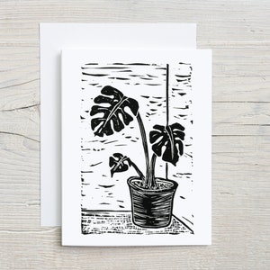 Puede incluir: Grabado en linóleo en blanco y negro de una planta de monstera en maceta con tres hojas. La planta está en una maceta texturizada y se coloca sobre un fondo de veta de madera.