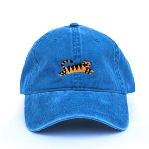 May include: A blue baseball cap with an embroidered tiger design. The tiger is orange and black, and the cap has a curved brim and adjustable strap. The cap is made of a soft, washed cotton material.