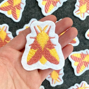 May include: A set of embroidered iron-on patches featuring a yellow and pink moth design. The patches are perfect for adding a touch of whimsy to clothing, bags, or other fabric items.