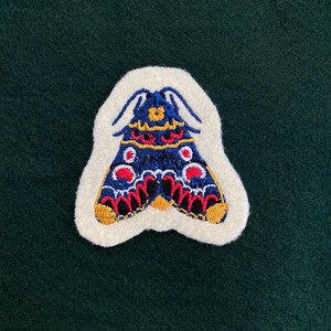 May include: A white felt patch with an embroidered blue, red, and gold butterfly. The butterfly has a detailed design with black outlines and white dots.