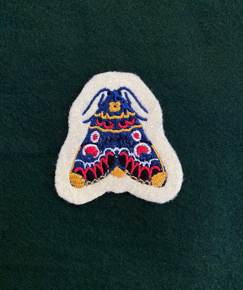 May include: A white felt patch with an embroidered blue, red, and gold butterfly. The butterfly has a detailed design with black outlines and white dots.