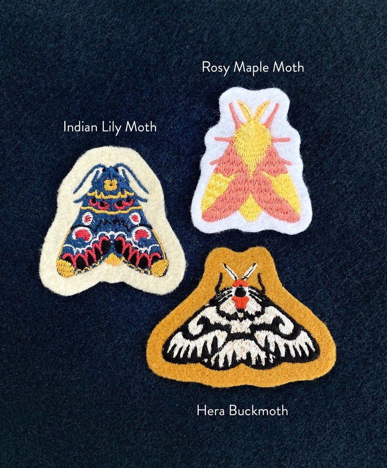 May include: Three embroidered moth patches on a dark blue fabric background. The patches are white with embroidered details in yellow, red, black, and blue. The patches are labeled "Rosy Maple Moth", "Indian Lily Moth", and "Hera Buckmoth".