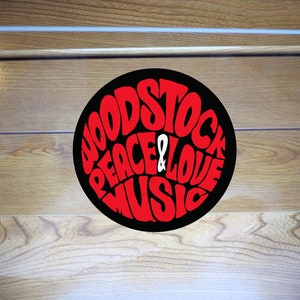 Könnte beinhalten: Ein klares, quadratisches Acrylschild mit den Worten "WOODSTOCK PEACE & LOVE MUSIC" in roter, stilisierter Schrift auf schwarzem, kreisförmigem Hintergrund. Das Schild befindet sich vor einem Holzgrund.
