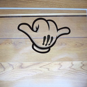 May include: A clear, rectangular acrylic sign featuring a black cartoon hand gesture, commonly known as the "hang loose" or "shaka" sign. The sign is set against a light brown wood grain background.