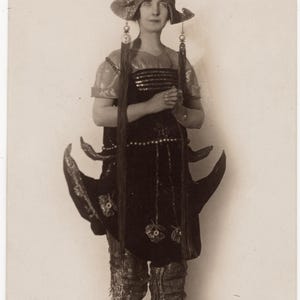 以下が含まれることがあります： 精巧な衣装を着た人物のヴィンテージセピア写真。衣装には、背の高い装飾的な頭飾り、暗く装飾されたチュニック、複雑なディテールのパンツが含まれています。長いタッセルが頭飾りとチュニックからぶら下がっています。靴はローヒールです。