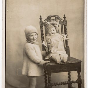 Può includere: Una fotografia vintage color seppia che ritrae un bambino con berretto, maglione e leggings in maglia in piedi accanto a una sedia di legno. Una bambola con un grande fiocco tra i capelli e un vestito chiaro è seduta sulla sedia intagliata.